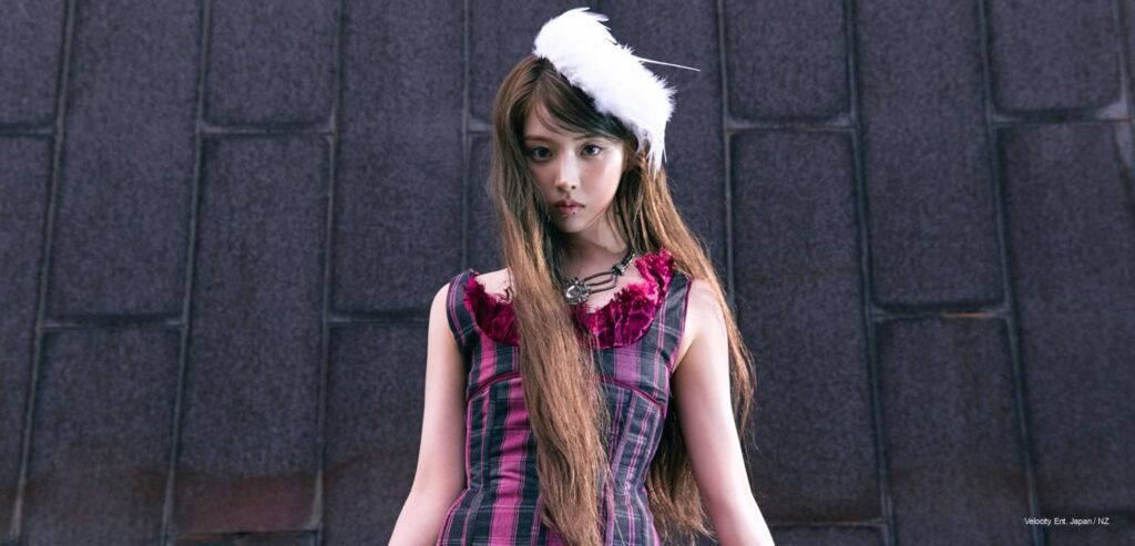 NMIXX member looking down at camera wearing pink tartan plaid corset dress and white feather headpiece against brutalist stone background.