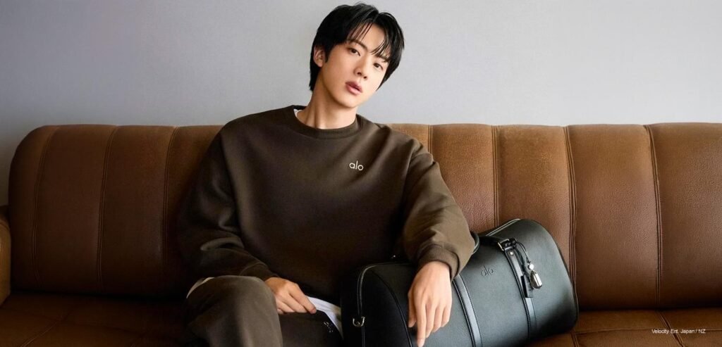 BTS Jin Esquire Wellness cover – Breaking K-Pop News BTS Jin posing for Esquire Wellness Korea x ALO Issue 03 cover in a black technical pullover hoodie and matching wide-leg cargo pants against a stone archway.
