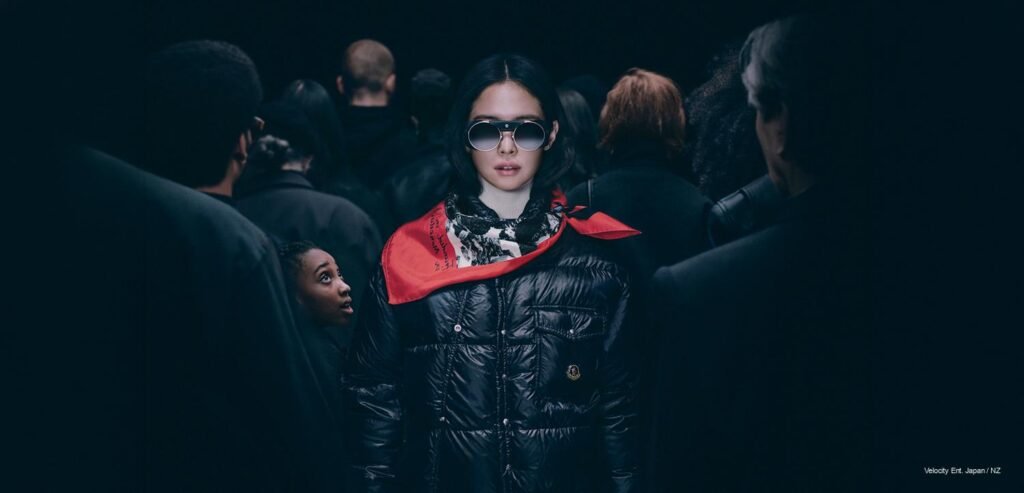 JENNIE ODD ATELIER fashion editorial BLACKPINK Jennie standing in a dark, atmospheric crowd wearing a black Moncler puffer jacket, tinted aviator goggles, and a red accent neck scarf for Moncler Re/Icons.
