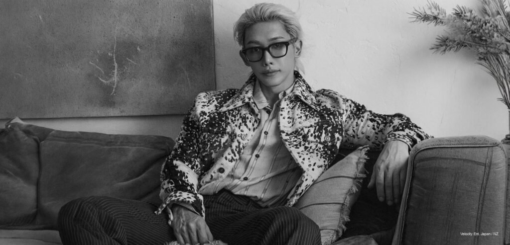 Wonho – Timid Magazine October 2026 Wonho in a studded patterned jacket, striped shirt, tinted sunglasses, and platinum hair sitting relaxed on a velvet sofa for Timid Magazine's October 2026 digital cover shoot.