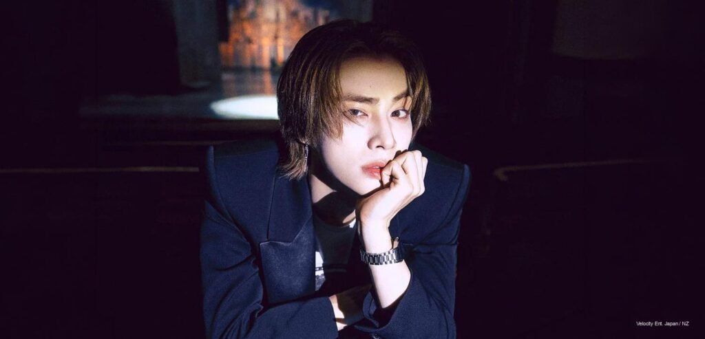 Xiaojun of WayV wearing a sharp navy suit leaning forward thoughtfully in a dramatic studio spotlight setting.